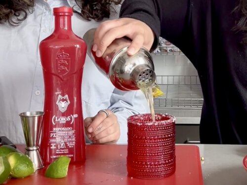 Hand pouring drink from cocktail shaker into red decorative glass